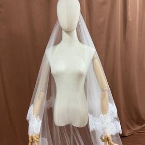 Puede incluir: Un velo de novia blanco con delicados bordes de encaje. El velo transparente se muestra sobre un maniqu&iacute;, destacando los intrincados detalles de encaje en los bordes. El fondo es de color marr&oacute;n.