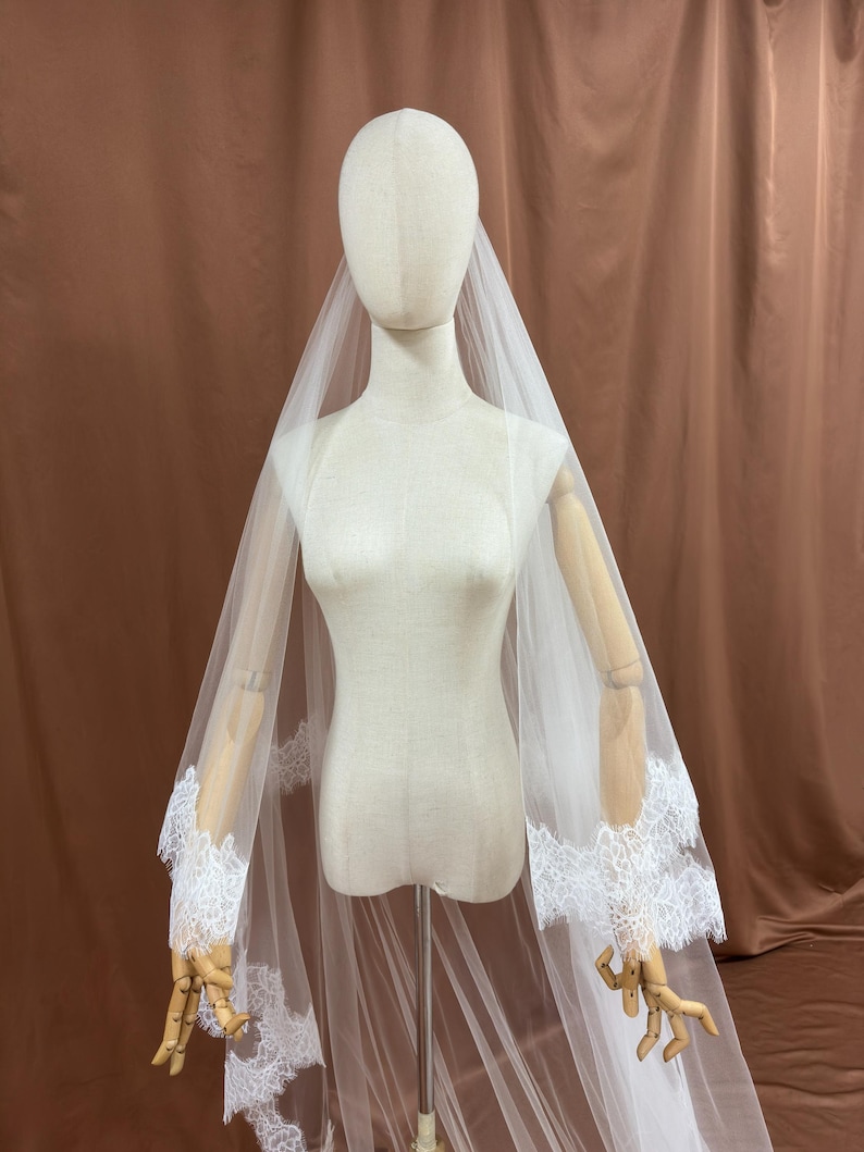 Puede incluir: Un velo de novia blanco con delicados bordes de encaje. El velo transparente se muestra sobre un maniqu&iacute;, destacando los intrincados detalles de encaje en los bordes. El fondo es de color marr&oacute;n.