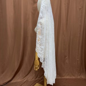 May include: A white lace bridal veil, draped over a mannequin head and shoulders. The veil is long, with a scalloped edge and intricate floral lace detailing. The mannequin is on a wooden stand against a brown backdrop.