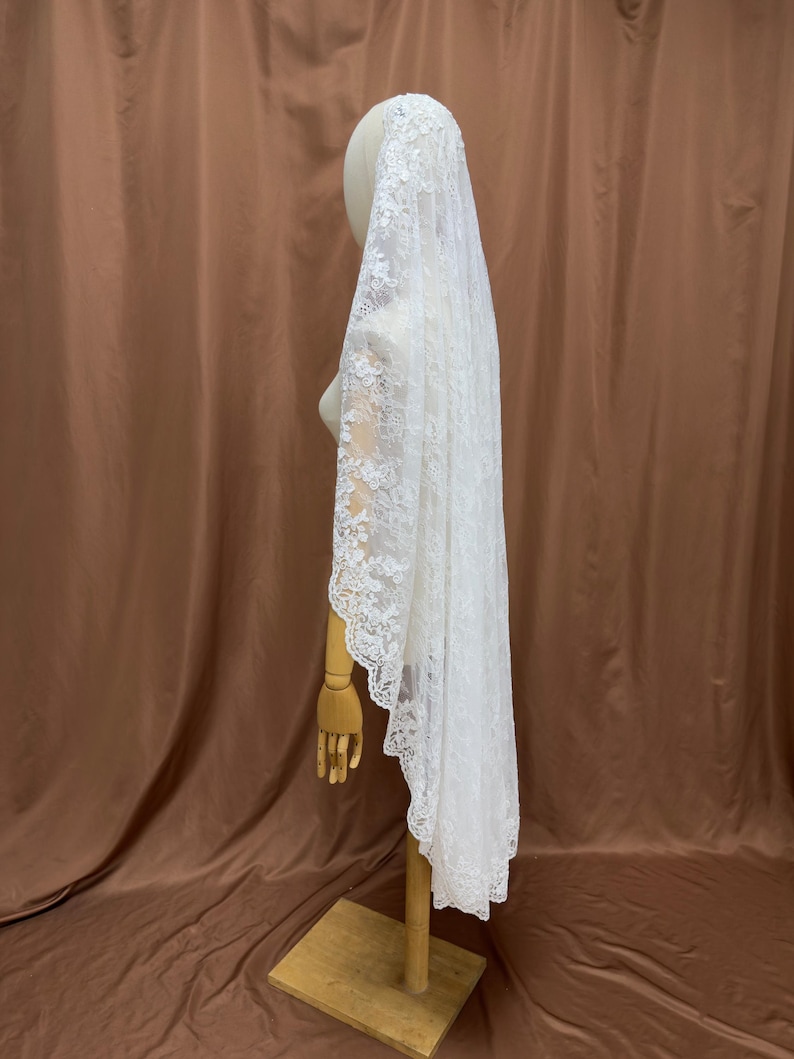 May include: A white lace bridal veil, draped over a mannequin head and shoulders. The veil is long, with a scalloped edge and intricate floral lace detailing. The mannequin is on a wooden stand against a brown backdrop.