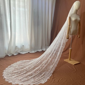 May include: A long, white bridal veil with intricate lace detailing along the edges. The veil is displayed on a mannequin, showcasing its flowing design. The background features white curtains and a brown backdrop.