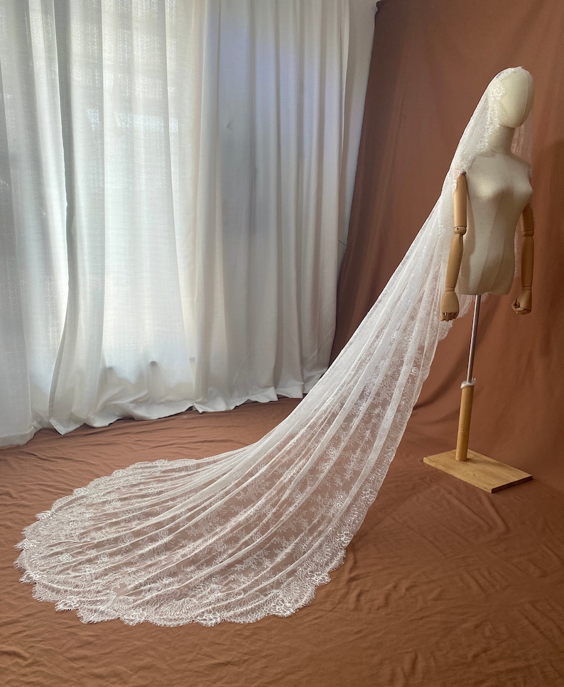 May include: A long, white bridal veil with intricate lace detailing along the edges. The veil is displayed on a mannequin, showcasing its flowing design. The background features white curtains and a brown backdrop.