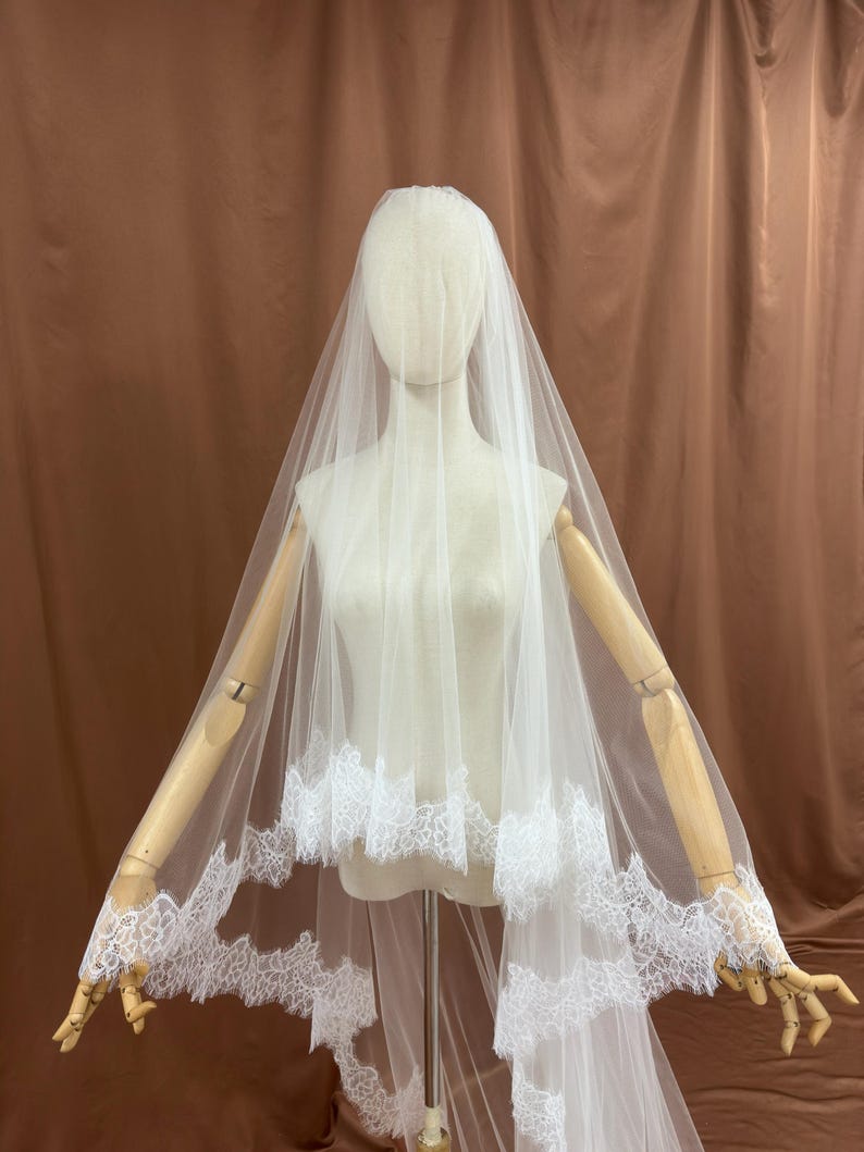 Puede incluir: Un velo de novia blanco con delicado ribete de encaje. El velo est&aacute; drapeado sobre un maniqu&iacute;, mostrando su dise&ntilde;o en capas y los intrincados detalles de encaje a lo largo de los bordes. El velo est&aacute; hecho de una tela transparente y ligera.