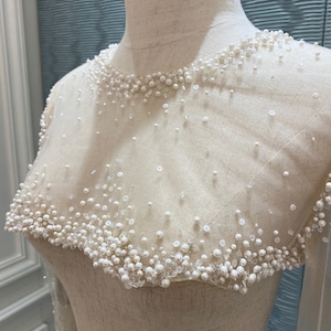 May include: Ivory-colored bridal gown bodice with a sheer overlay, adorned with scattered pearls and sequins. The neckline and shoulders are heavily embellished with clustered pearls, creating a delicate, textured effect. Long sleeves are visible.