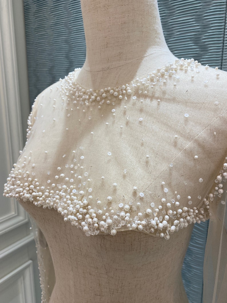 May include: Ivory-colored bridal gown bodice with a sheer overlay, adorned with scattered pearls and sequins. The neckline and shoulders are heavily embellished with clustered pearls, creating a delicate, textured effect. Long sleeves are visible.