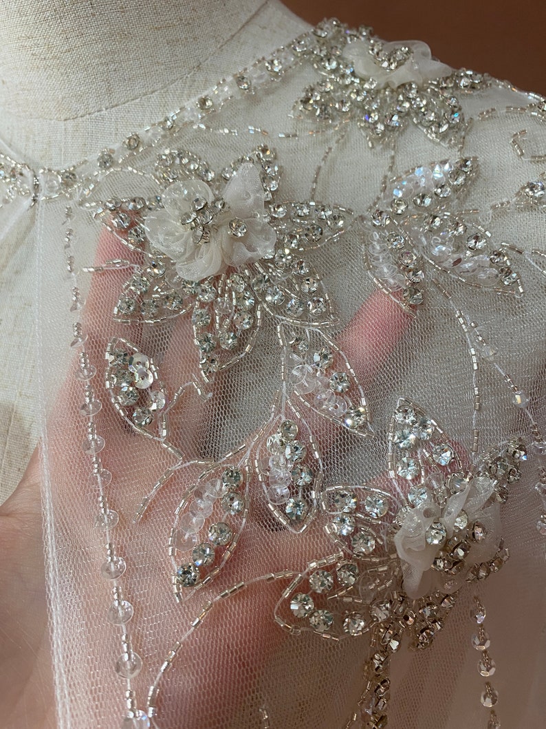 Puede incluir: Primer plano de una tela blanca y transparente adornada con brillantes pedrer&iacute;a y cuentas. Los dise&ntilde;os florales y de hojas se crean con la pedrer&iacute;a, y peque&ntilde;as cuentas delinean los bordes. La tela es probablemente para ropa de novia o formal.