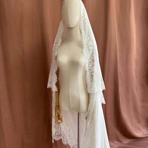 Puede incluir: Un velo de novia blanco con intrincados bordes de encaje est&aacute; drapeado sobre un maniqu&iacute;. El velo cae por la espalda, mostrando los delicados detalles del encaje. El maniqu&iacute; est&aacute; sobre un soporte de madera, sobre un fondo neutro.