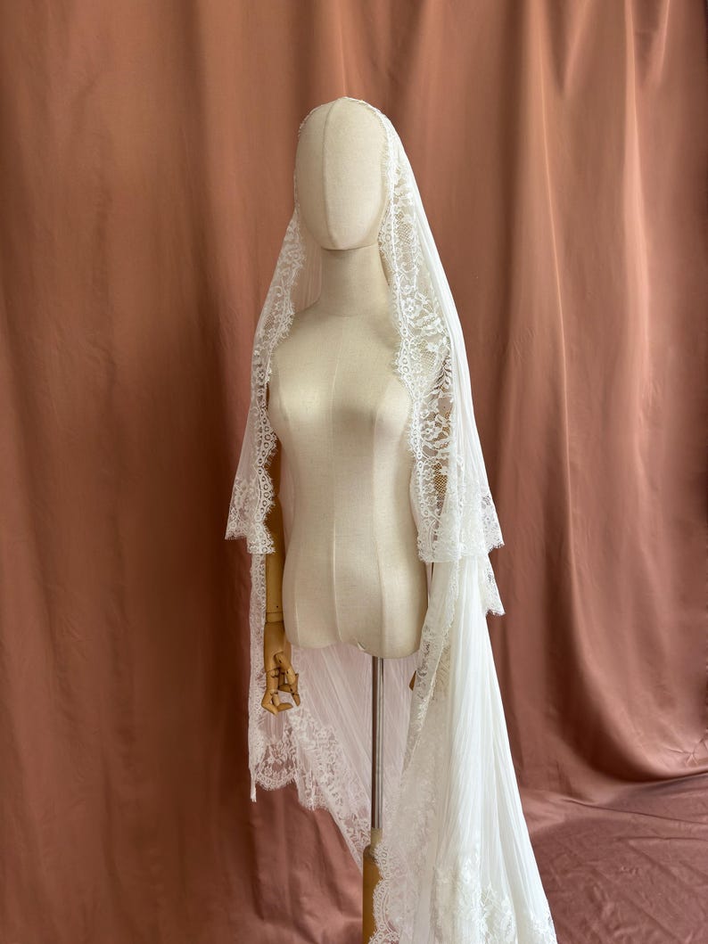 Puede incluir: Un velo de novia blanco con intrincados bordes de encaje est&aacute; drapeado sobre un maniqu&iacute;. El velo cae por la espalda, mostrando los delicados detalles del encaje. El maniqu&iacute; est&aacute; sobre un soporte de madera, sobre un fondo neutro.