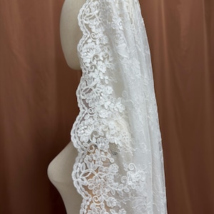 May include: A white lace veil with a floral design. The veil is draped over a mannequin head, showcasing the intricate details of the lace. The edges of the veil are adorned with a scalloped floral pattern.