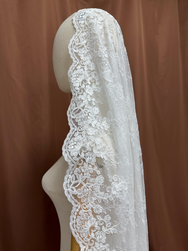 May include: A white lace veil with a floral design. The veil is draped over a mannequin head, showcasing the intricate details of the lace. The edges of the veil are adorned with a scalloped floral pattern.
