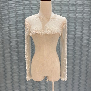May include: A sheer, long-sleeved top adorned with small, white pearls. The neckline and cuffs are heavily embellished with pearls, creating a delicate, textured effect. The top is displayed on a mannequin.