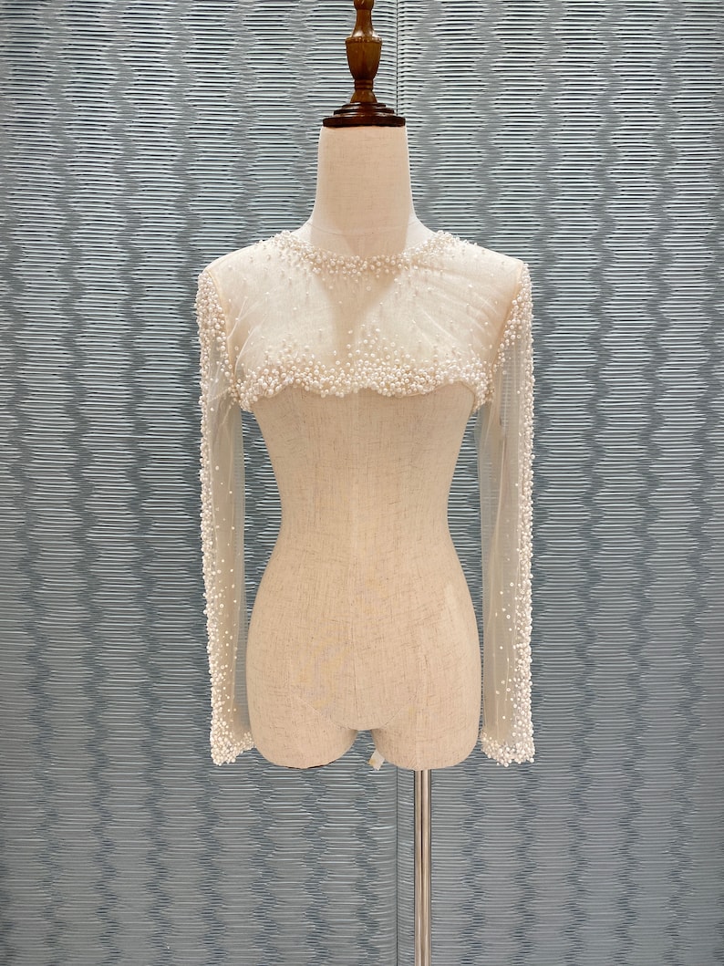 May include: A sheer, long-sleeved top adorned with small, white pearls. The neckline and cuffs are heavily embellished with pearls, creating a delicate, textured effect. The top is displayed on a mannequin.