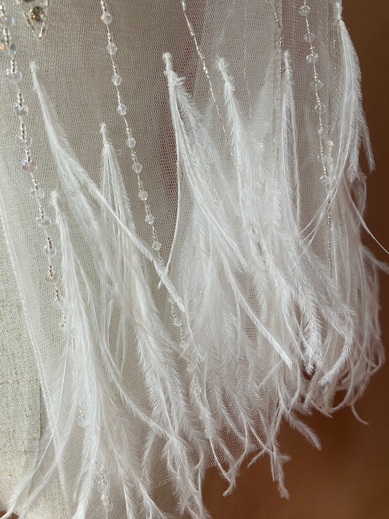 Puede incluir: Primer plano de una tela blanca transparente adornada con delicadas plumas blancas y hebras de cuentas transparentes. Las plumas caen en cascada, creando un efecto suave y texturizado. Las cuentas a&ntilde;aden un toque de brillo al dise&ntilde;o.