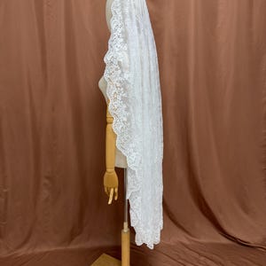 May include: A white lace veil with a scalloped edge is displayed on a wooden mannequin. The veil is long and flowing, with intricate floral patterns along the border. The background is a solid brown color.
