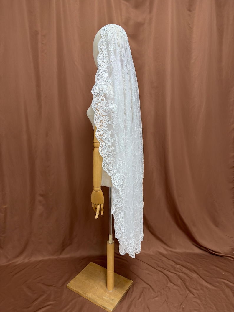 May include: A white lace veil with a scalloped edge is displayed on a wooden mannequin. The veil is long and flowing, with intricate floral patterns along the border. The background is a solid brown color.