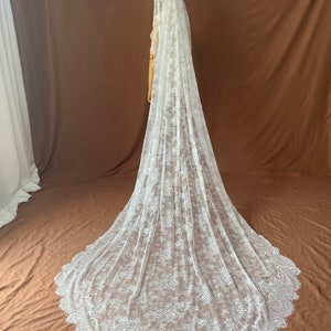 May include: A long, white lace wedding veil with a scalloped edge. The veil is draped over a mannequin, showcasing its intricate floral pattern and flowing design. The veil is made of delicate, sheer fabric.