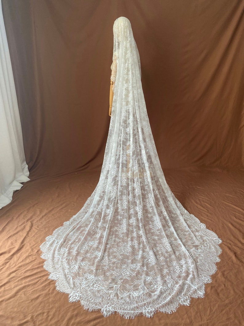 May include: A long, white lace wedding veil with a scalloped edge. The veil is draped over a mannequin, showcasing its intricate floral pattern and flowing design. The veil is made of delicate, sheer fabric.