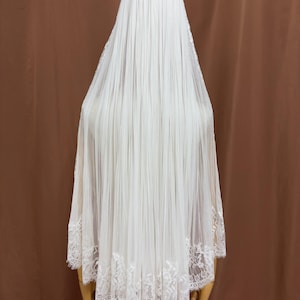 Puede incluir: Un velo de novia blanco con un dise&ntilde;o plisado. El velo est&aacute; hecho de una tela transparente y presenta un delicado ribete de encaje a lo largo del borde inferior. El velo est&aacute; drapeado sobre un maniqu&iacute;.