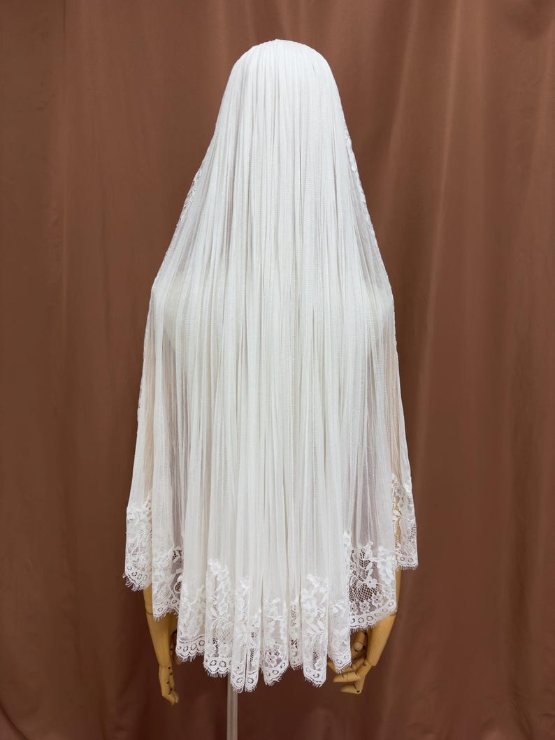 Puede incluir: Un velo de novia blanco con un dise&ntilde;o plisado. El velo est&aacute; hecho de una tela transparente y presenta un delicado ribete de encaje a lo largo del borde inferior. El velo est&aacute; drapeado sobre un maniqu&iacute;.