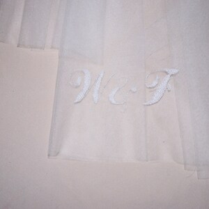 May include: Close-up of a sheer, white fabric with embroidered initials "W-F" in a delicate script. The fabric appears to be layered, creating a soft, ethereal effect. The background is a pale, neutral color.