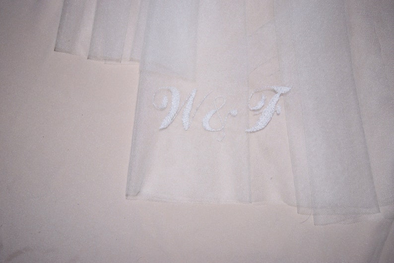 May include: Close-up of a sheer, white fabric with embroidered initials "W-F" in a delicate script. The fabric appears to be layered, creating a soft, ethereal effect. The background is a pale, neutral color.