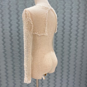 May include: A sheer, off-white garment with long sleeves and a collar, adorned with small, white beads. The garment is displayed on a mannequin, showcasing its delicate design and texture. The sleeves are long and fitted, and the collar is square.