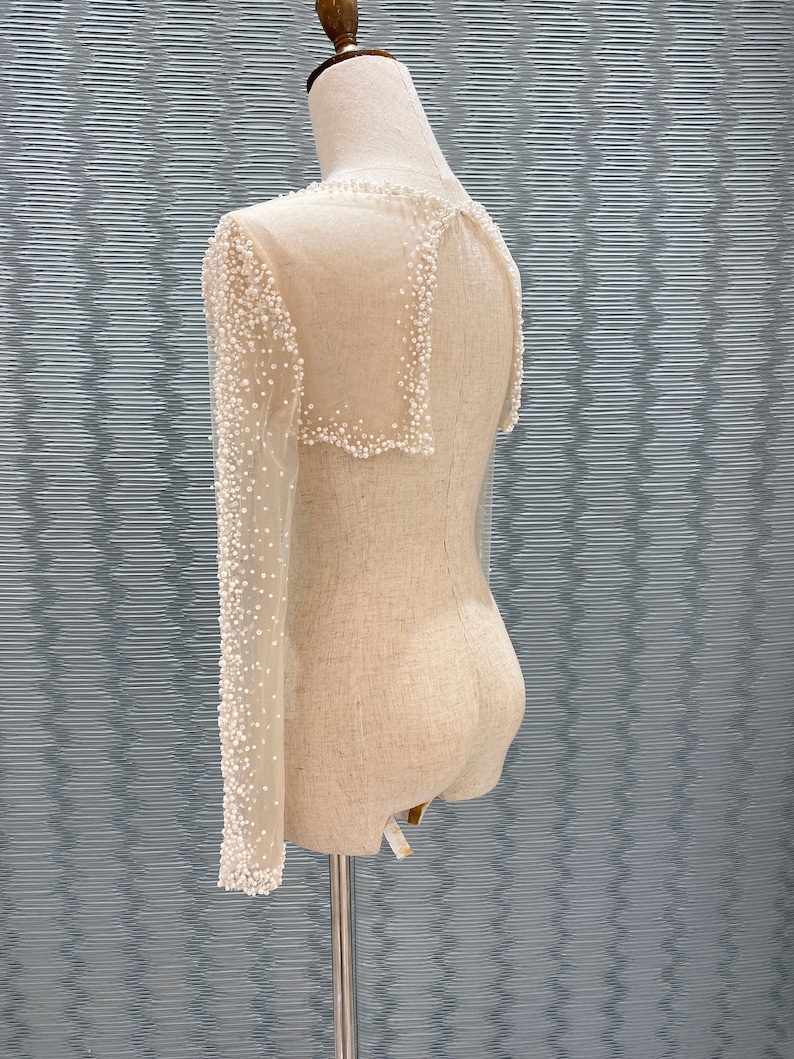 May include: A sheer, off-white garment with long sleeves and a collar, adorned with small, white beads. The garment is displayed on a mannequin, showcasing its delicate design and texture. The sleeves are long and fitted, and the collar is square.