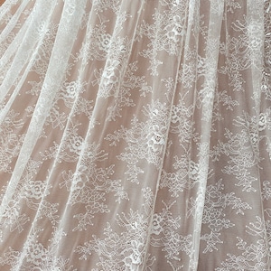 May include: Close-up of white lace fabric with a floral pattern. The delicate lace is draped, showing the intricate details of the design. The fabric is semi-transparent, revealing the surface beneath. Ideal for bridal wear or decorative projects.