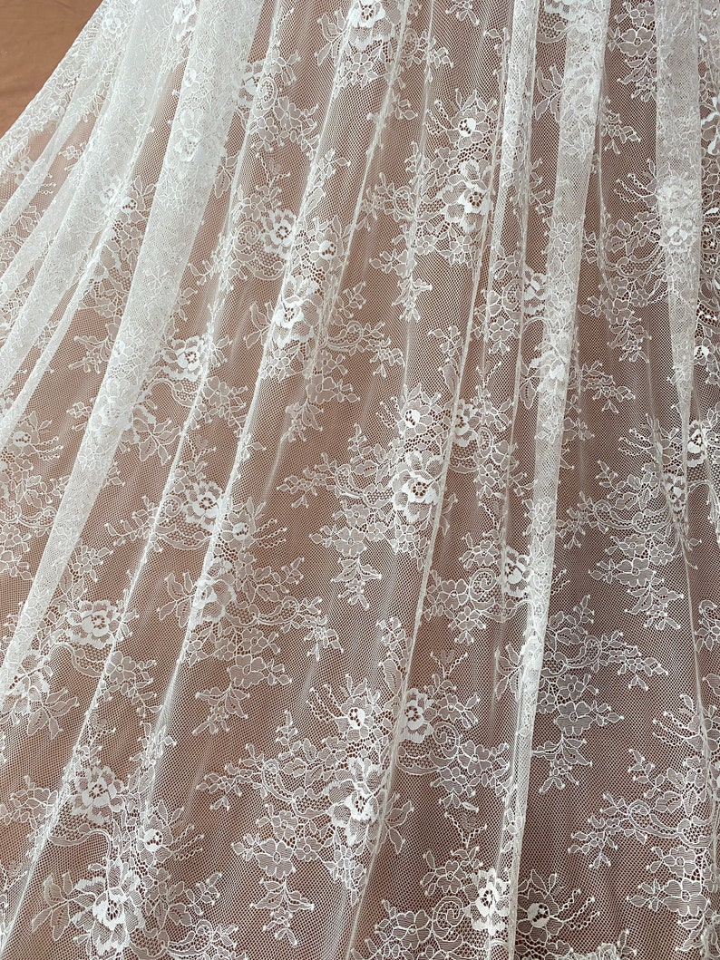 May include: Close-up of white lace fabric with a floral pattern. The delicate lace is draped, showing the intricate details of the design. The fabric is semi-transparent, revealing the surface beneath. Ideal for bridal wear or decorative projects.