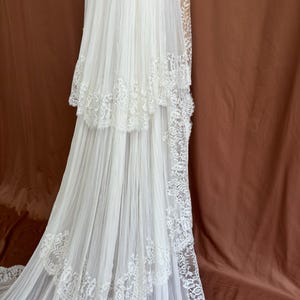 Puede incluir: Un vestido de novia blanco con una falda escalonada y una larga cola. El vestido presenta delicados adornos de encaje a lo largo de los bordes de los niveles y la cola. La tela parece ligera y fluida, creando un aspecto rom&aacute;ntico y et&eacute;reo.