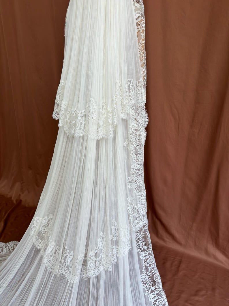 Puede incluir: Un vestido de novia blanco con una falda escalonada y una larga cola. El vestido presenta delicados adornos de encaje a lo largo de los bordes de los niveles y la cola. La tela parece ligera y fluida, creando un aspecto rom&aacute;ntico y et&eacute;reo.