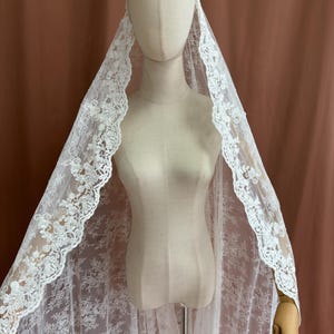 May include: A white lace veil with floral detailing is draped over a mannequin. The veil's edges are scalloped, and the lace pattern is visible throughout. The mannequin is on a stand, and the background is a solid brown color.