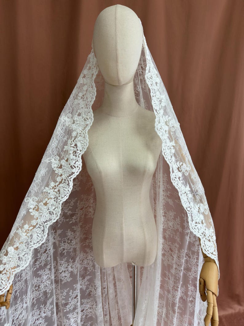 May include: A white lace veil with floral detailing is draped over a mannequin. The veil's edges are scalloped, and the lace pattern is visible throughout. The mannequin is on a stand, and the background is a solid brown color.