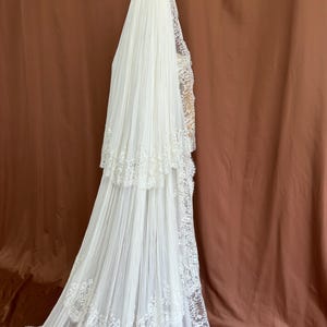 Puede incluir: Un velo de novia blanco con un dise&ntilde;o en capas y un intrincado ribete de encaje. El velo cae en cascada, creando un efecto fluido. El fondo es de color marr&oacute;n liso.
