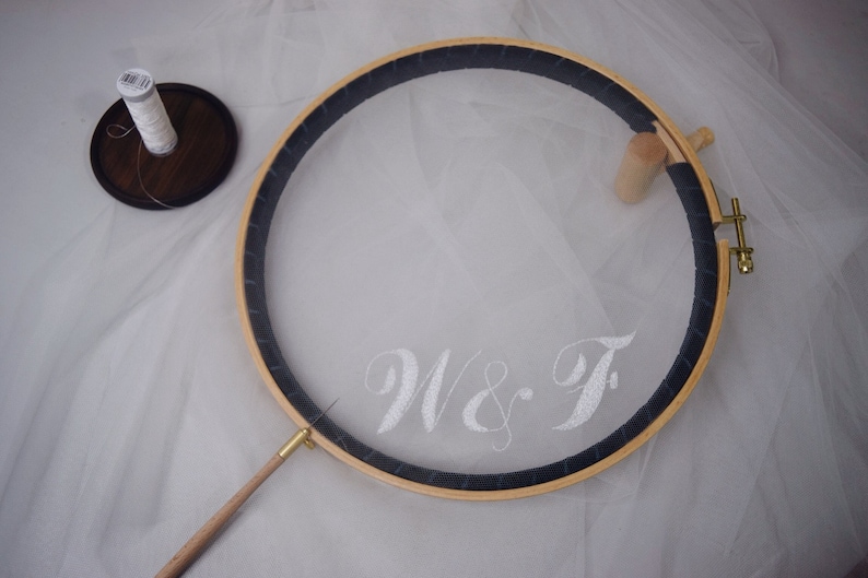 May include: A wooden embroidery hoop with white fabric stretched inside, featuring the embroidered initials "W & F". A spool of white thread sits on a small wooden disc. The hoop has a dark blue border.