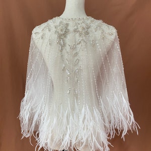 Puede incluir: Una capa de novia blanca y transparente con intrincados abalorios y adornos florales alrededor del escote. La capa presenta l&iacute;neas verticales con cuentas y est&aacute; rematada con una franja de plumas blancas en el dobladillo. La capa se exhibe en un maniqu&iacute;.
