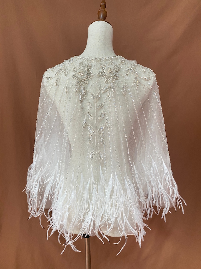 Puede incluir: Una capa de novia blanca y transparente con intrincados abalorios y adornos florales alrededor del escote. La capa presenta l&iacute;neas verticales con cuentas y est&aacute; rematada con una franja de plumas blancas en el dobladillo. La capa se exhibe en un maniqu&iacute;.