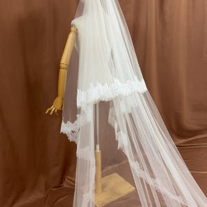 Puede incluir: Un velo de novia blanco con delicados bordes de encaje. El velo se exhibe en un maniqu&iacute;, mostrando su dise&ntilde;o en capas y su tejido fluido. Los detalles de encaje a&ntilde;aden un toque de elegancia al accesorio de boda.