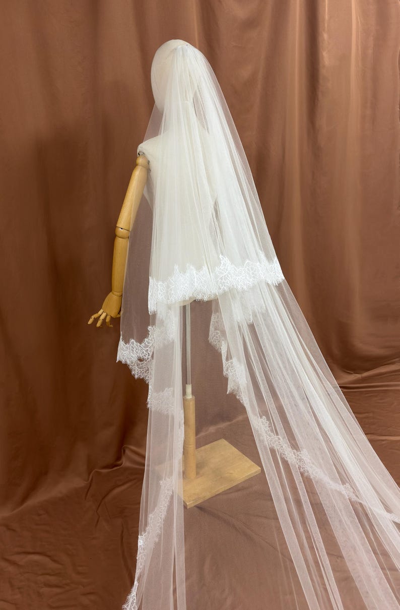 Puede incluir: Un velo de novia blanco con delicados bordes de encaje. El velo se exhibe en un maniqu&iacute;, mostrando su dise&ntilde;o en capas y su tejido fluido. Los detalles de encaje a&ntilde;aden un toque de elegancia al accesorio de boda.