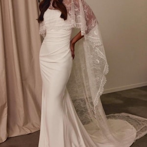 Puede incluir: Un vestido blanco largo hasta el suelo con corpiño ajustado y cintura fruncida, combinado con una capa de encaje transparente que llega al suelo, con intrincados patrones florales y bordes festoneados que caen sobre los hombros formando una cola.