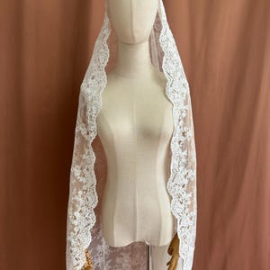 May include: A white lace veil with a floral pattern and scalloped edges. The veil is draped over a mannequin, showcasing its delicate design and texture. The background is a solid, muted brown.