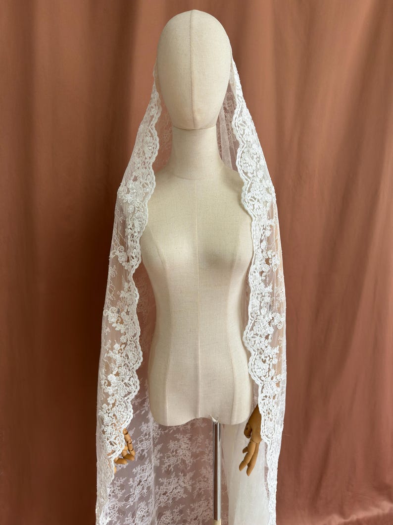 May include: A white lace veil with a floral pattern and scalloped edges. The veil is draped over a mannequin, showcasing its delicate design and texture. The background is a solid, muted brown.