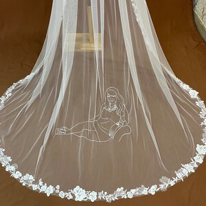 May include: A long, white bridal veil with a floral lace trim. The veil features a delicate illustration of a person reclining on a chair. The veil is made of sheer fabric and is displayed against a brown background.