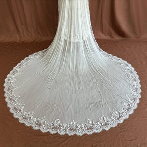 Puede incluir: Un velo de novia blanco con una larga cola fluida. El velo est&aacute; hecho de una tela transparente con un dise&ntilde;o plisado y un delicado y ornamentado ribete de encaje a lo largo de los bordes. La cola se extiende sobre un fondo marr&oacute;n.