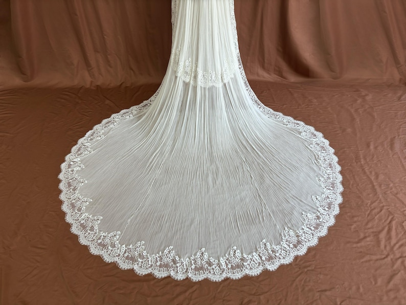 Puede incluir: Un velo de novia blanco con una larga cola fluida. El velo est&aacute; hecho de una tela transparente con un dise&ntilde;o plisado y un delicado y ornamentado ribete de encaje a lo largo de los bordes. La cola se extiende sobre un fondo marr&oacute;n.