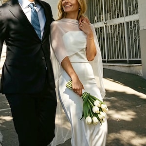 Puede incluir: Un vestido largo de satén blanco con una capa de organza transparente, acompañado de un ramo de tulipanes blancos. El conjunto se completa con sandalias blancas de tacón ancho, logrando un estilo monocromático de texturas elegantes.
