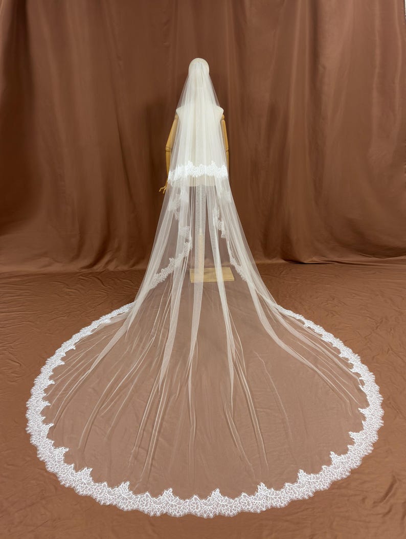 Puede incluir: Un velo de novia blanco largo con un delicado ribete de encaje. El velo se exhibe en un maniqu&iacute;, mostrando su longitud y su intrincado dise&ntilde;o. El ribete de encaje a&ntilde;ade un toque de elegancia a la tela transparente.
