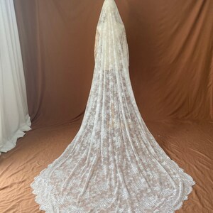 May include: A long, white lace wedding veil with a floral pattern. The veil is draped over a mannequin, showcasing its length and intricate design. The veil's edge has a scalloped trim.
