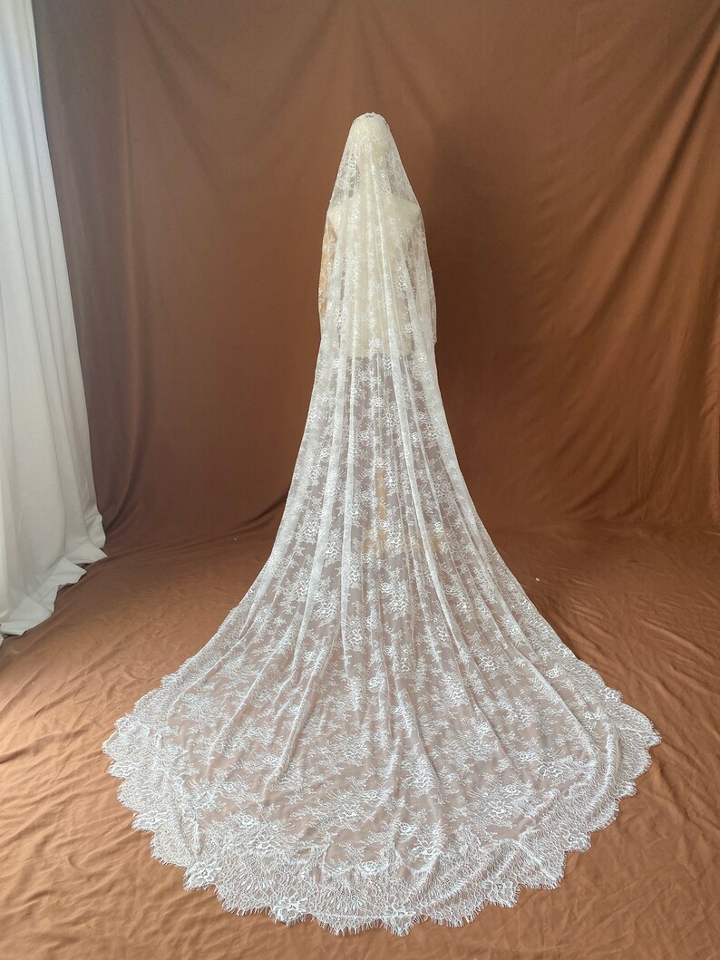 May include: A long, white lace wedding veil with a floral pattern. The veil is draped over a mannequin, showcasing its length and intricate design. The veil's edge has a scalloped trim.