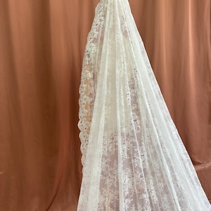 May include: A long, white lace veil with intricate floral patterns. The veil is draped over a mannequin, showcasing its delicate design and texture. The background is a solid, muted orange-brown.